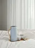 A light blue thermos with a wooden handle sits on a tray with a cup of coffee, an egg, and a slice of toast, all on a white, rumpled bed. Produktbild-Vorschau