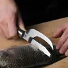 A person is using a fish scaler tool to remove scales from a fish on a wooden cutting board. Náhled obrázku produktu