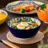 A table setting with various dishes and bowls, including a salad in a decorative blue bowl. Produktbild-Vorschau