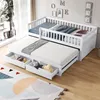 A white wooden daybed with a pull-out trundle bed and storage drawers, set up in a bright room with a large window and a colorful rug. Produktbild-Vorschau