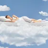 A woman is sleeping on a mattress that appears to be floating on clouds in a blue sky. Anteprima dell’immagine prodotto