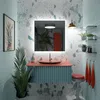 A modern bathroom vanity with a pink countertop and teal cabinet, featuring a round glass sink, a large LED-lit mirror, and decorative elements like plants and towels. Produktbild-Vorschau