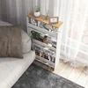 A narrow white shelving unit with a wooden top sits next to a sofa, filled with various items like jars, plants, and books. Voir les images du produit