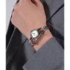 A close-up shot of a woman's wrist wearing a silver Guess watch with a white face and black accents, layered over a grey blazer. Produktbild-Vorschau