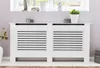 A white radiator cover with horizontal slats is shown in a room with a window in the background. Podgląd zdjęcia produktu
