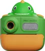 A green dinosaur-shaped instant print camera with orange accents, shown from the front against a white background. Voir les images du produit