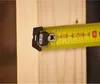 A close-up of a yellow tape measure with black markings and a black end hook, resting against a light-colored wooden surface. Voir les images du produit