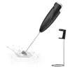 A black electric milk frother with a silver whisk attachment is shown in a splash of milk, with its black and silver stand to the right. The background is plain white. Produktbild-Vorschau