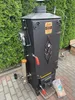 A black electric smoker is set up outdoors on a brick patio, with a green hedge in the background. Náhled obrázku produktu