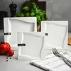 A set of white square plates with a wavy design, arranged on a table with a tomato and a fork. Náhled obrázku produktu