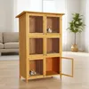 A wooden multi-level rabbit hutch with open doors, placed indoors on a wooden floor with a sofa and a plant in the background. Produktbild-Vorschau