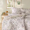 A bedroom scene with a bed dressed in floral patterned bedding, a bedside table with a lamp and plant, and framed botanical prints on the wall. Produktbild-Vorschau