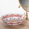 A decorative ceramic sink with a colorful floral pattern is placed on a wooden countertop, with a brass faucet and a mirror in the background. Náhled obrázku produktu