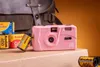 A pink Kodak camera is displayed with film rolls and a backpack in the background. Produktbild-Vorschau