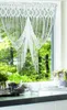 A white lace curtain with fringe hangs in a window, with green trees and foliage visible outside. Plants and decorative items are on the windowsill. Produktbild-Vorschau