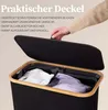 A hand is opening the lid of a black laundry basket with a wooden rim. Inside, there are two compartments with white and purple laundry. Produktbild-Vorschau