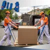 Two movers are using lifting straps to move a large cardboard box on a pallet towards a van. The straps are worn by the movers and attached to the box. The top left corner shows a close-up of the blue lifting straps. Produktbild-Vorschau