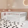A white wooden children's bed with a gate is shown in a room with cloud decorations on the walls and a patterned rug on the floor. Produktbild-Vorschau