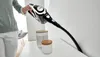 A person is using a Bosch handheld vacuum cleaner to clean a narrow space between a wall and a surface. There are two decorative jars on the surface. Produktbild-Vorschau
