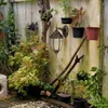 An outdoor wall lamp is mounted on a textured wall, surrounded by various potted plants and garden elements. Produktbild-Vorschau