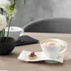 A white coffee cup with a frothy beverage sits on a white saucer with a small pastry on the side, next to a vase of white flowers. Produktbild-Vorschau