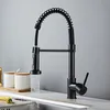 A black kitchen faucet with a pull-down sprayer is shown in use, with water flowing into a sink. Two white jars with wooden lids are visible on the counter behind the faucet. Produktbild-Vorschau