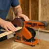 A person is using an orange belt sander to sand a piece of wood. Voir les images du produit