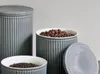 Three grey ceramic canisters with ribbed detailing, two filled with coffee beans and tea leaves respectively, and one lid is placed next to a canister. Produktbild-Vorschau