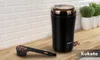 A black electric coffee grinder with a transparent lid is placed on a wooden countertop, next to a scoop filled with coffee beans. The background features white subway tiles and potted plants. Podgląd zdjęcia produktu