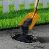 A shovel pouring black asphalt into a hole in a paved surface, with green grass and a white picket fence in the background. Produktbild-Vorschau