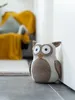 A stuffed owl doorstop is placed on a floor next to a wall, with a blurred background of a chair and a yellow blanket. Anteprima dell’immagine prodotto