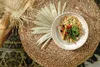 A bowl of food is placed on a woven surface, with a dried palm leaf and a fork nearby. Produktbild-Vorschau
