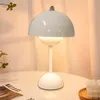 A white mushroom-shaped table lamp is placed on a white table next to a book and a candle. Produktbild-Vorschau