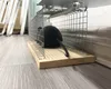 A black toy mouse is inside a metal wire cage trap, which is placed on a wooden base on a wooden floor. Produktbild-Vorschau