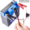 A hand is holding a battery charger connector near a car battery. Produktbild-Vorschau