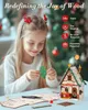 A young girl is assembling a wooden Christmas house model, with the finished product and assembly instructions visible on the table. Produktbild-Vorschau