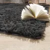 A close-up view of a dark gray shaggy rug with an open book resting on it, placed on a light wooden floor. Produktbild-Vorschau