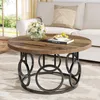 A round coffee table with a wood-grain top and a black metal base with circular accents, placed in a living room setting with a sofa and a window in the background. Podgląd zdjęcia produktu