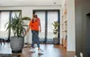 A woman is tending to a large potted plant in a modern living room. Náhľad obrázku produktu