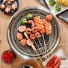A top-down view of a BBQ grill with various food items on skewers, placed on a wooden table. Produktbild-Vorschau