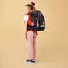 A young girl wearing a backpack and casual clothing, posing against a plain background. Náhled obrázku produktu
