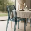 A transparent chair is placed at a dining table set for a meal, with a window showing greenery in the background. Produktbild-Vorschau