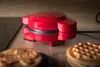 A red mini waffle maker is placed on a wooden surface, with waffles in the foreground. Produktbild-Vorschau