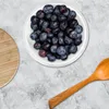 A top-down view of a white bowl filled with blueberries, placed on a grey marble-like surface. A wooden cutting board is on the left, and a wooden spoon is on the right. Produktbild-Vorschau