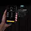 A hand holding a smartphone displaying a Godox app, with a camera and flash unit in the background. Náhľad obrázku produktu