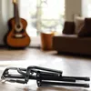 A folded guitar stand is in the foreground, with a guitar and furniture in the background. Náhľad obrázku produktu