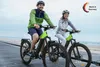A man and a woman are riding electric mountain bikes on a wooden boardwalk near the water. Produktbild-Vorschau