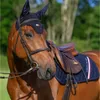 A close-up of a brown horse wearing a saddle and bridle, with a navy blue saddle pad and ear bonnet. Produktbild-Vorschau