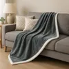 A gray blanket with a white sherpa trim is draped over a gray sofa, with two decorative pillows behind it. A lamp and a small plant are visible on a side table to the left. Produktbild-Vorschau