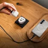 A hand is plugging a white adapter into a black adapter on a wooden table, with a smartphone charging nearby. Produktbild-Vorschau